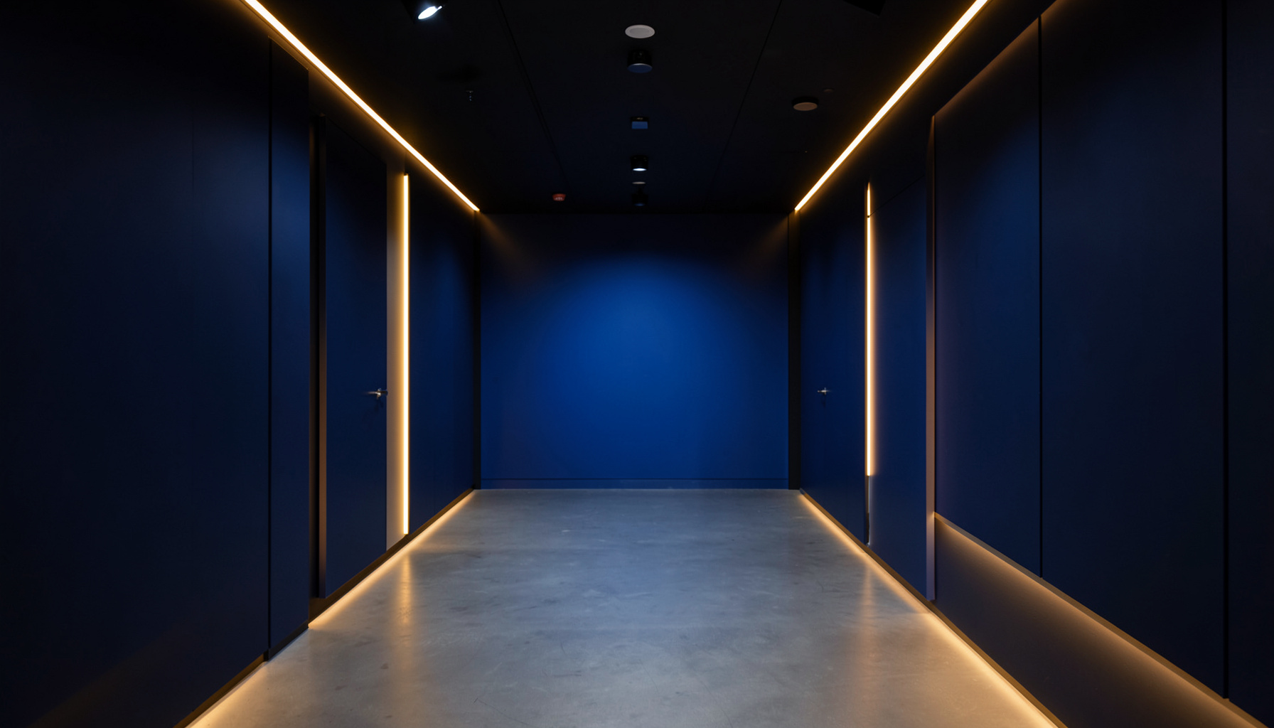 A symmetrical wide-angle view of a modern television studio corridor with clean lines, deep navy walls, and warm amber accent lighting, conveying a calm broadcast atmosphere.